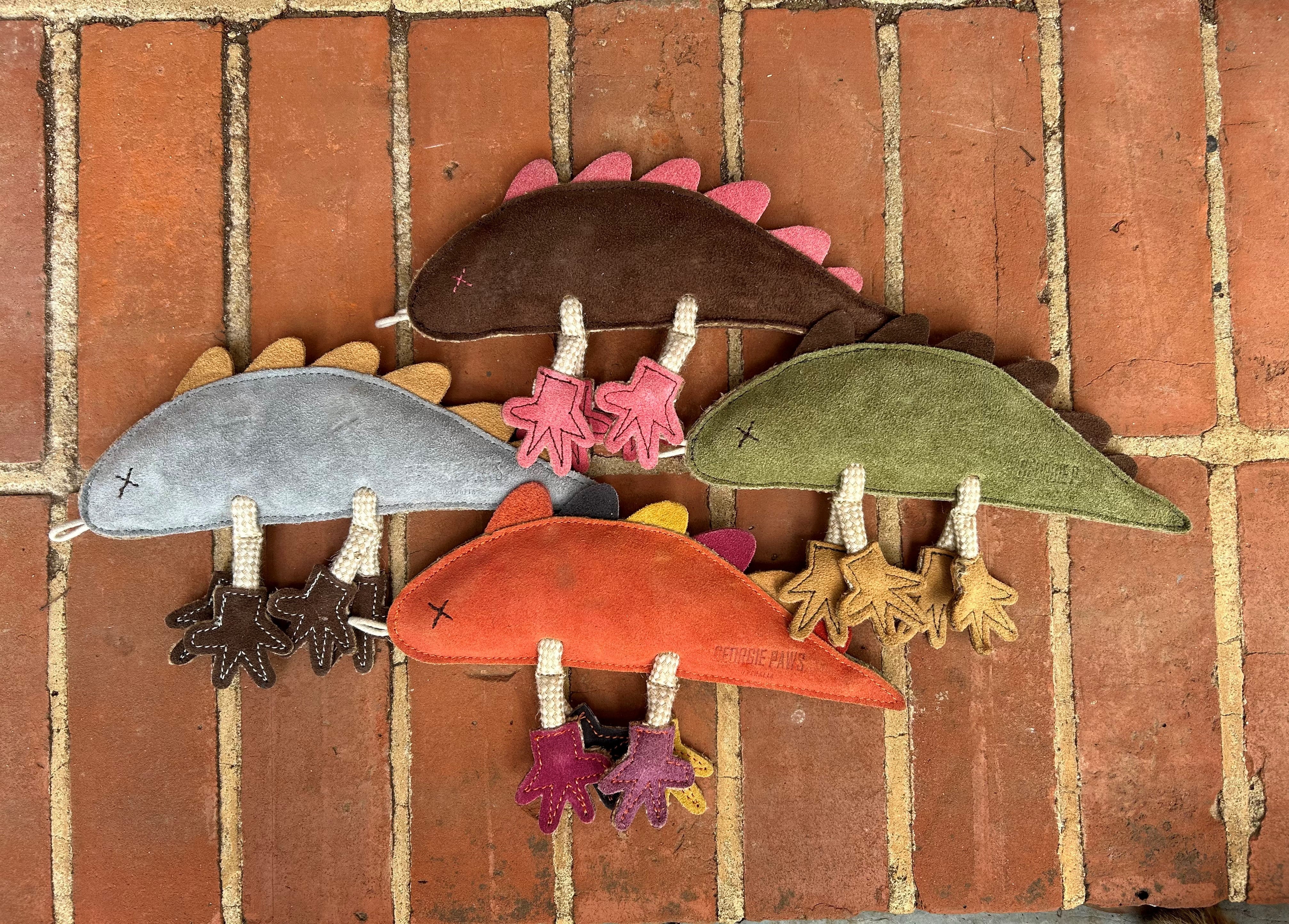 Four handmade plush dinosaurs with different colored bodies and spikes (gray/yellow, brown/pink, red/yellow, green/pink) are displayed on a brick surface. Each has stitched legs and a small cross-stitched "X" for eyes.