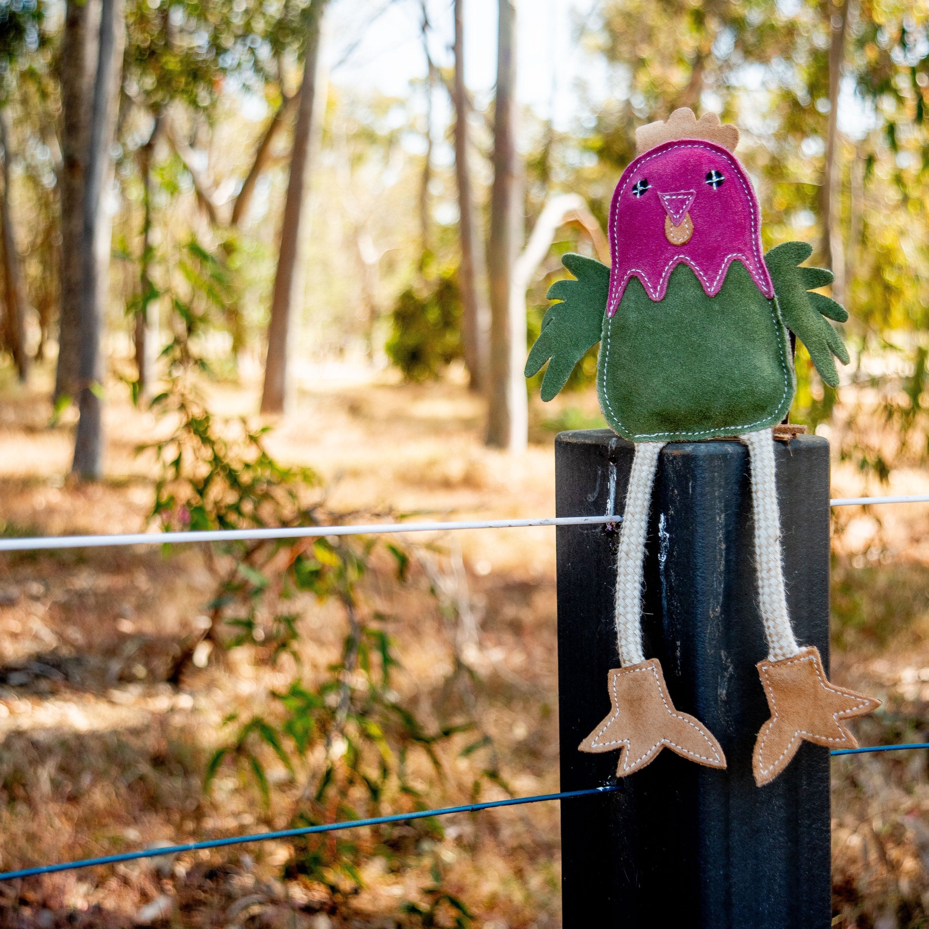Matilda the Chicken - Chive, a vibrant creation by Georgie Paws, features sustainable Buffalo Suede and is perched on a black wooden post. The toy showcases a pink head, green body, and yellow beak, with beige legs that dangle gracefully. It sits outdoors among trees and dry grass under bright sunlight against a softly blurred background.