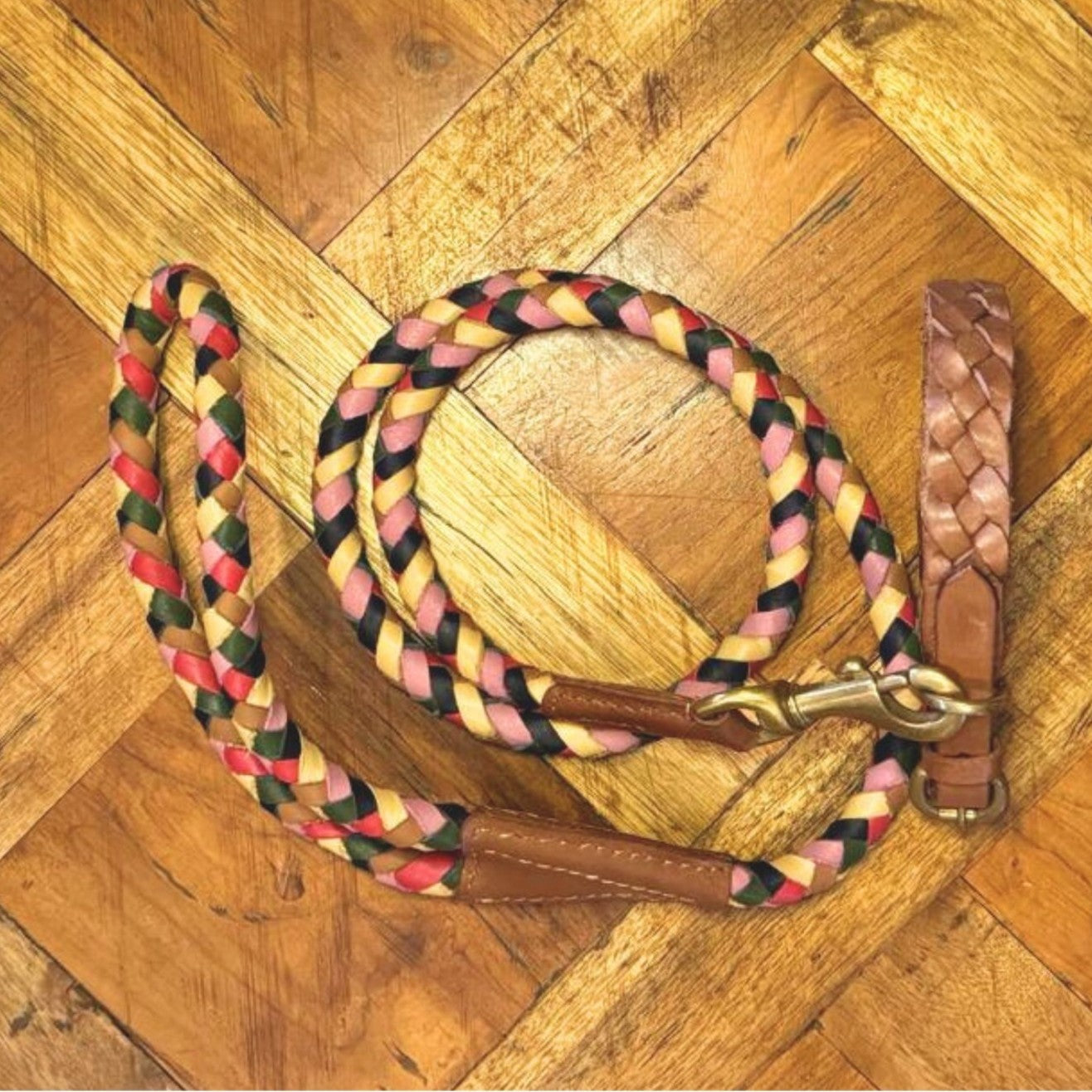 A braided dog leash with red, yellow, black, and tan strands lies coiled on a wooden floor. It features leather accents and brass hardware, ready to attach to the Georgie Paws Casey Collar - Syrup.