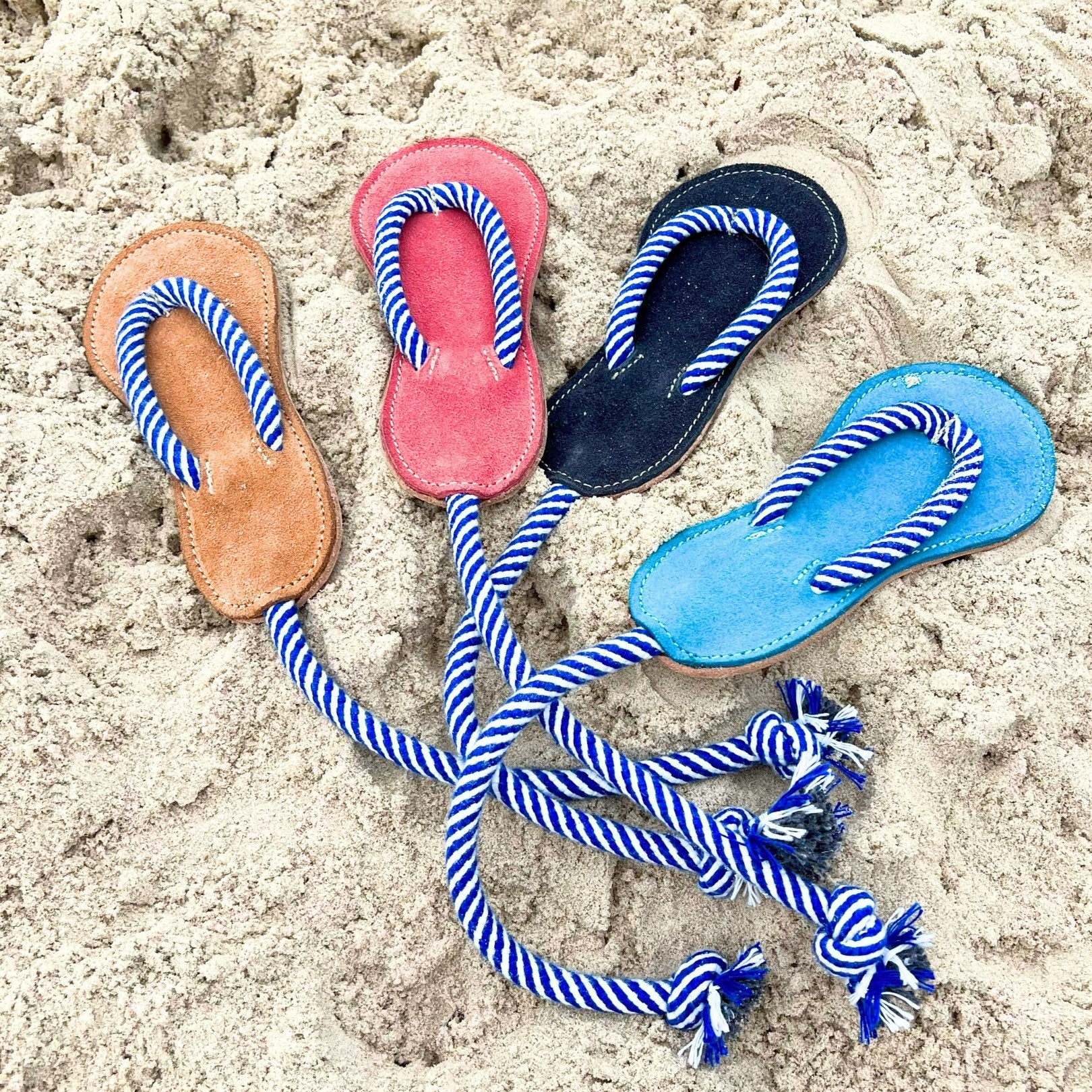 Three pairs of Georgie Paws pink Jandals with blue and white striped straps are arranged in a triangle on sandy beach terrain, suggesting a relaxed day at the sea with puppies playing nearby.