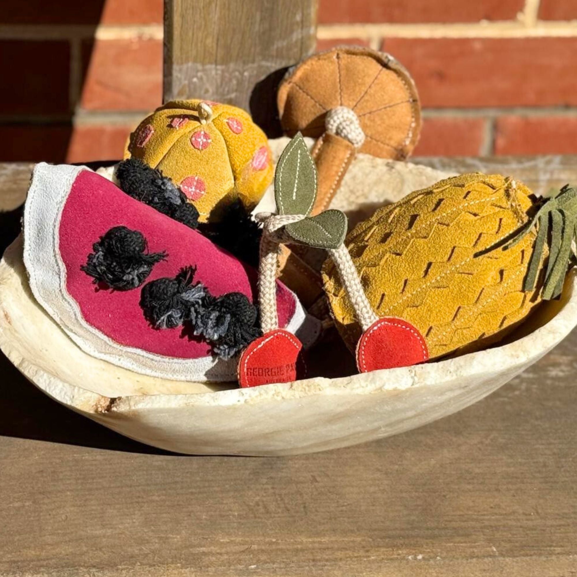 A wooden bowl holds plush toys shaped like a watermelon slice, pineapple, cherries, mushroom, and an orange. They are brightly colored and textured, sitting on a wooden surface with a brick wall backdrop.