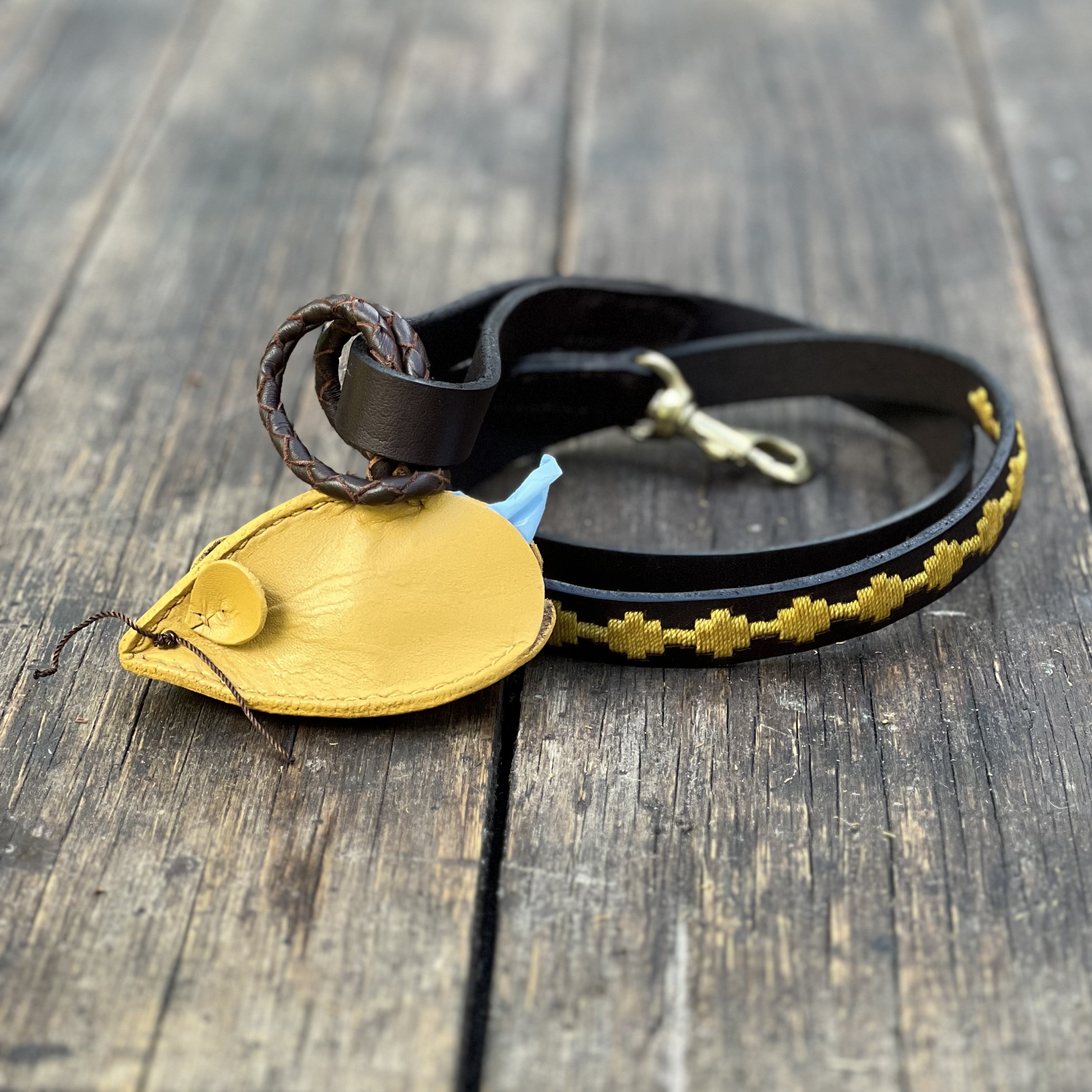 A black leather dog leash with a brass clasp, featuring a yellow leather pouch and decorative yellow stitching, is coiled on a rustic wooden surface.