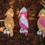 Three vibrant, handmade dog toys named Gav the Galah from Georgie Paws rest on a brown, earthy ground. The left toy is tan, the middle one is pink with white and gray details, and the right toy showcases pink with green accents. Each features stitched eyes and knotted string legs. Small twigs and leaves are scattered around them.