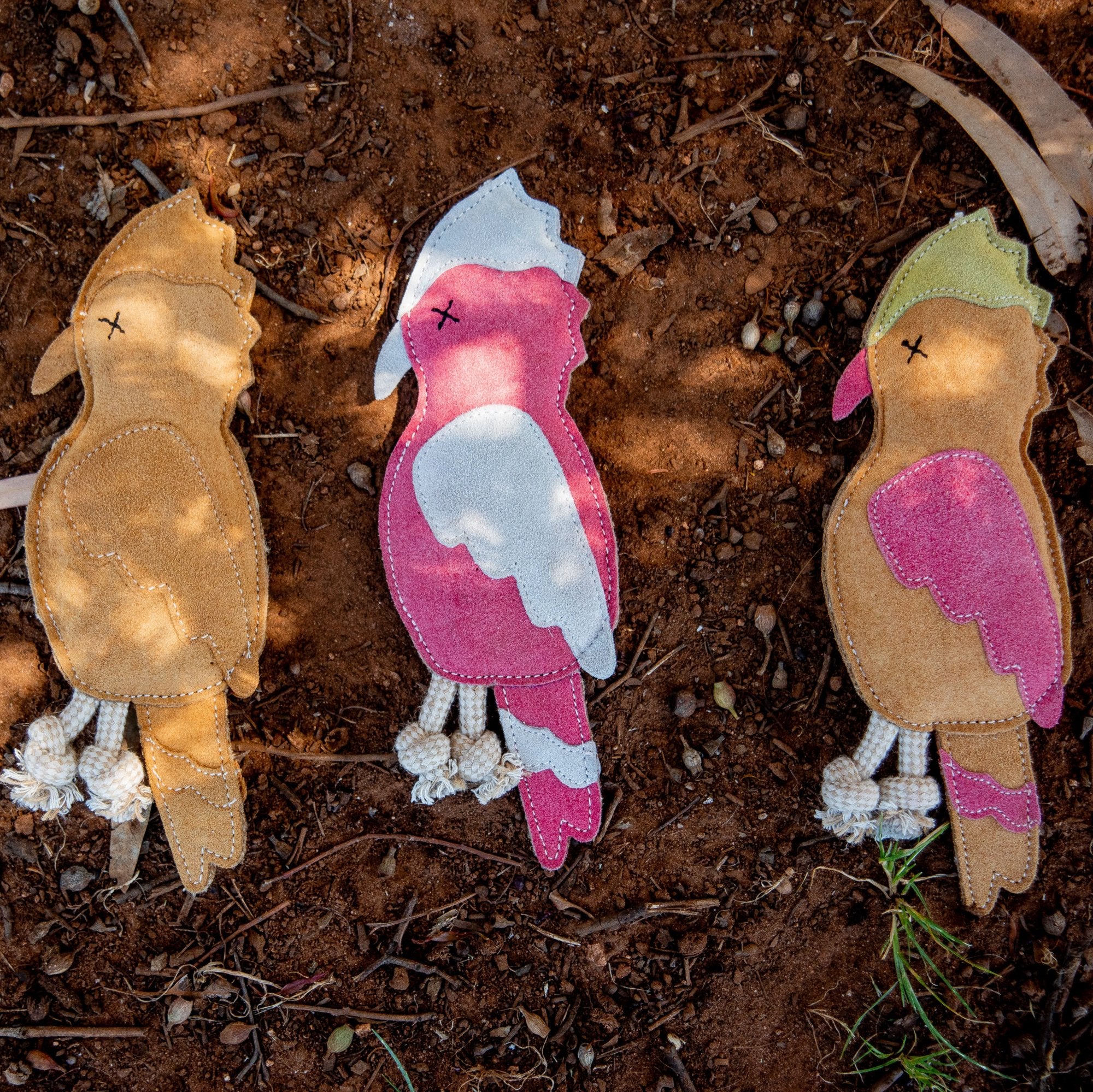 Three vibrant, handmade dog toys named Gav the Galah from Georgie Paws rest on a brown, earthy ground. The left toy is tan, the middle one is pink with white and gray details, and the right toy showcases pink with green accents. Each features stitched eyes and knotted string legs. Small twigs and leaves are scattered around them.