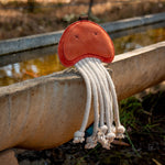 A whimsical toy from Georgie Paws, the Joe Jellyfish - ochre, features a bright ochre body with long, white, rope-like tentacles that hangs playfully over the edge of a concrete water trough. The sunny outdoor setting highlights its playful design and casts shadows on this compostable dog toy.