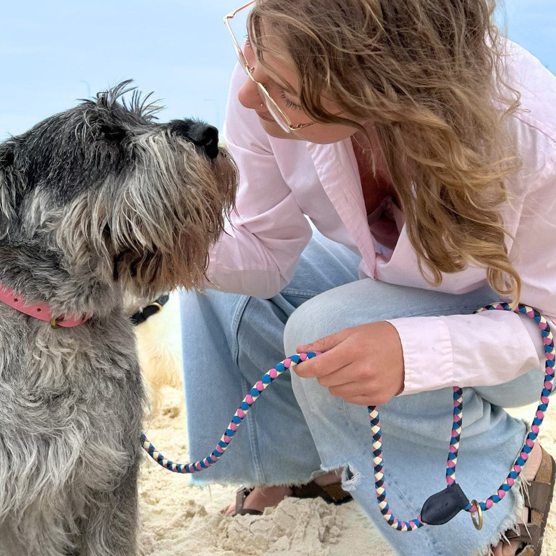 A woman in glasses and a light pink shirt kneels on sand, holding the Georgie Paws Beach Lead attached to a large shaggy gray dog. They share a warm, affectionate moment together at the beach.