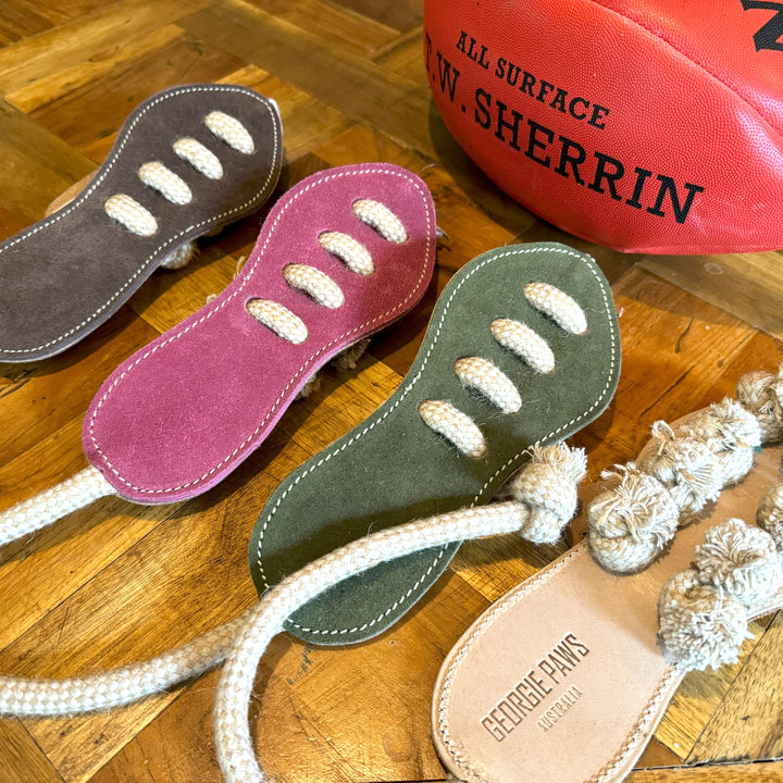 Three Georgie Paws "Footy Boot - Chive" eco-friendly dog toys in sandal shapes (brown, pink, green) with white rope straps and pom-poms rest on wood beside a red Sherrin football; one sole reads "GEORGE PAWS AUSTRALIA.