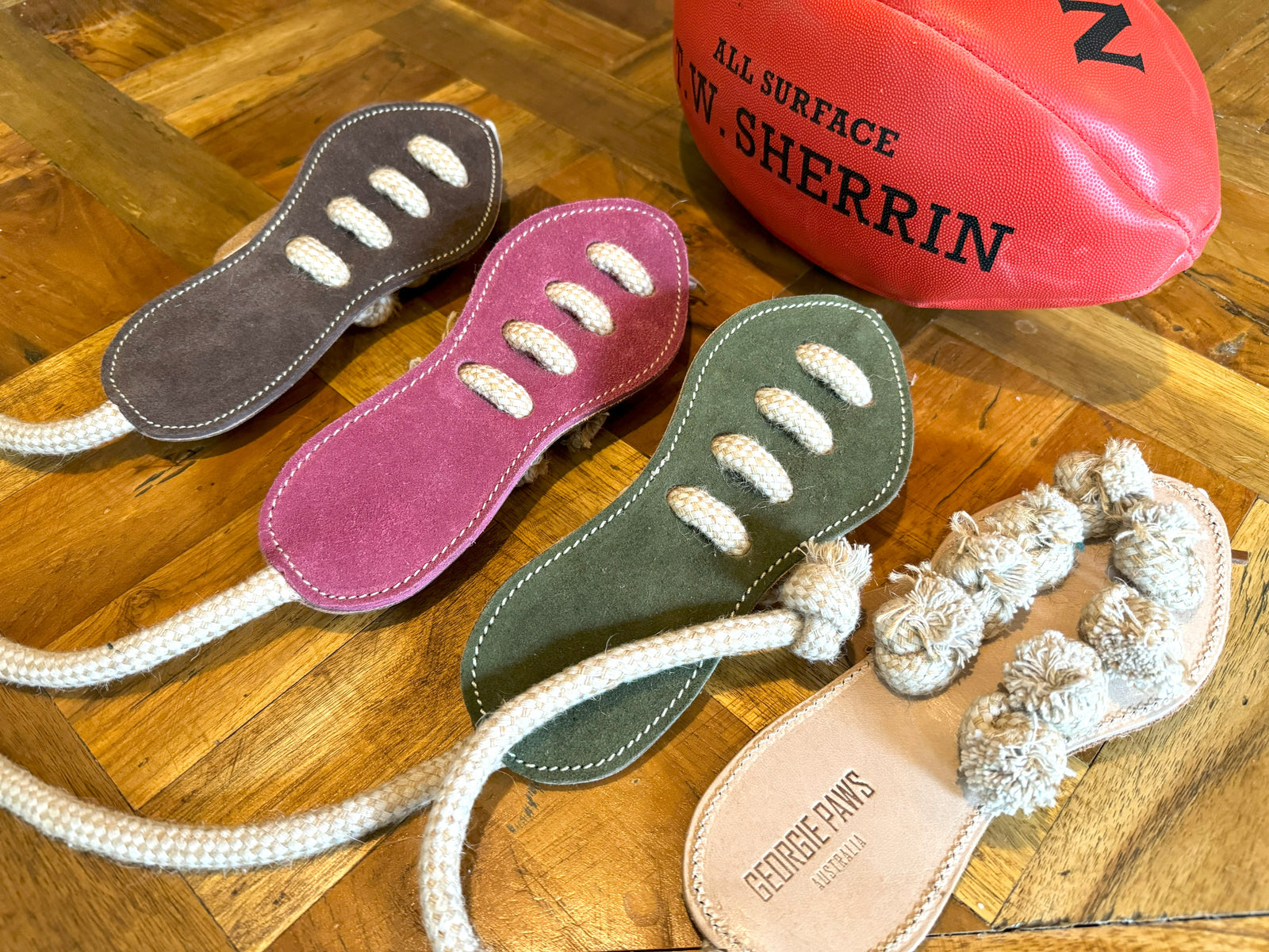 Three Footy Boot sandals in brown, green, and hot pink by Georgie Paws with thick rope straps are displayed on wood next to a red Sherrin football. The hot pink eco-friendly sandal features intricate knotted rope detailing on top.