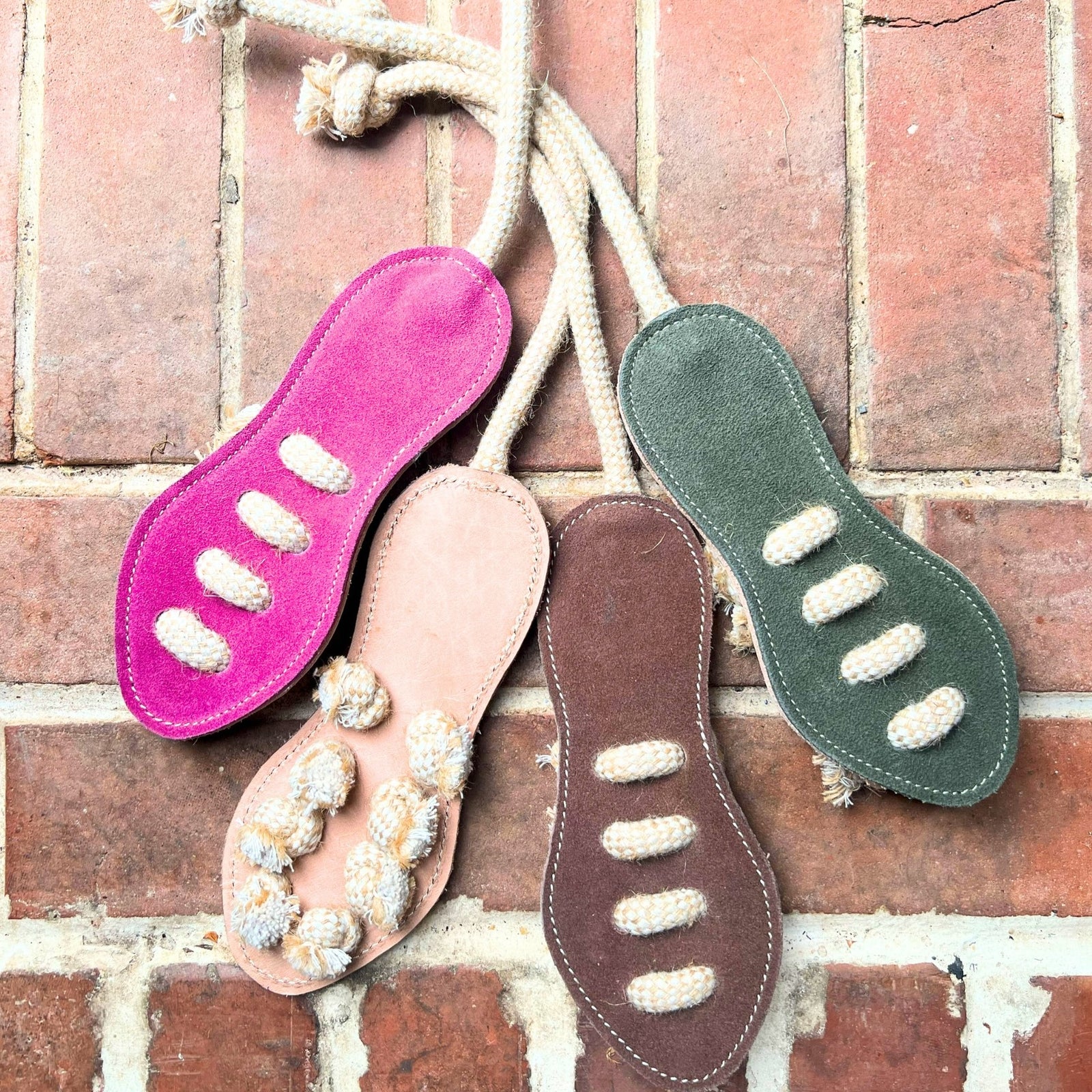 Four Georgie Paws Footy Boot - raw dog tug toys made from eco-friendly buffalo leather in pink, tan, brown, and green lie on red bricks. Each has three center-threaded rope sections and a top rope loop for tugging fun.