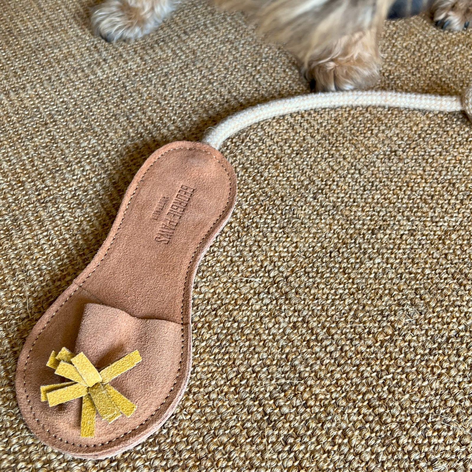 The Georgie Paws Sigrid Slipper - natural, a tan sandal-shaped Buffalo Leather dog toy with yellow tassels and rope, lies on a woven rug. A small fluffy dog's paws and lower legs are visible at the top of the image.