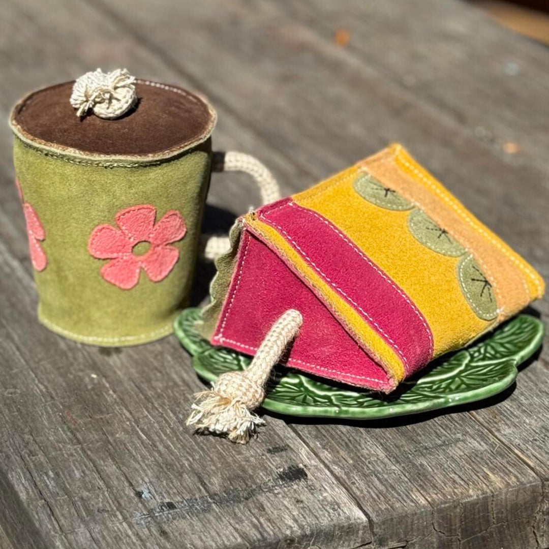A styled photo of the Hotto Chocco Dog Toy by Georgie Paws next to a slice of cake, a plush toy shaped like a mug of hot chocolate.