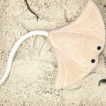 A pink Simon the Stingray - chalk kite with black eyes and a long white tail made from coconut fibre rests on coarse sand, evoking thoughts of marine life as it blends into its beachy backdrop.