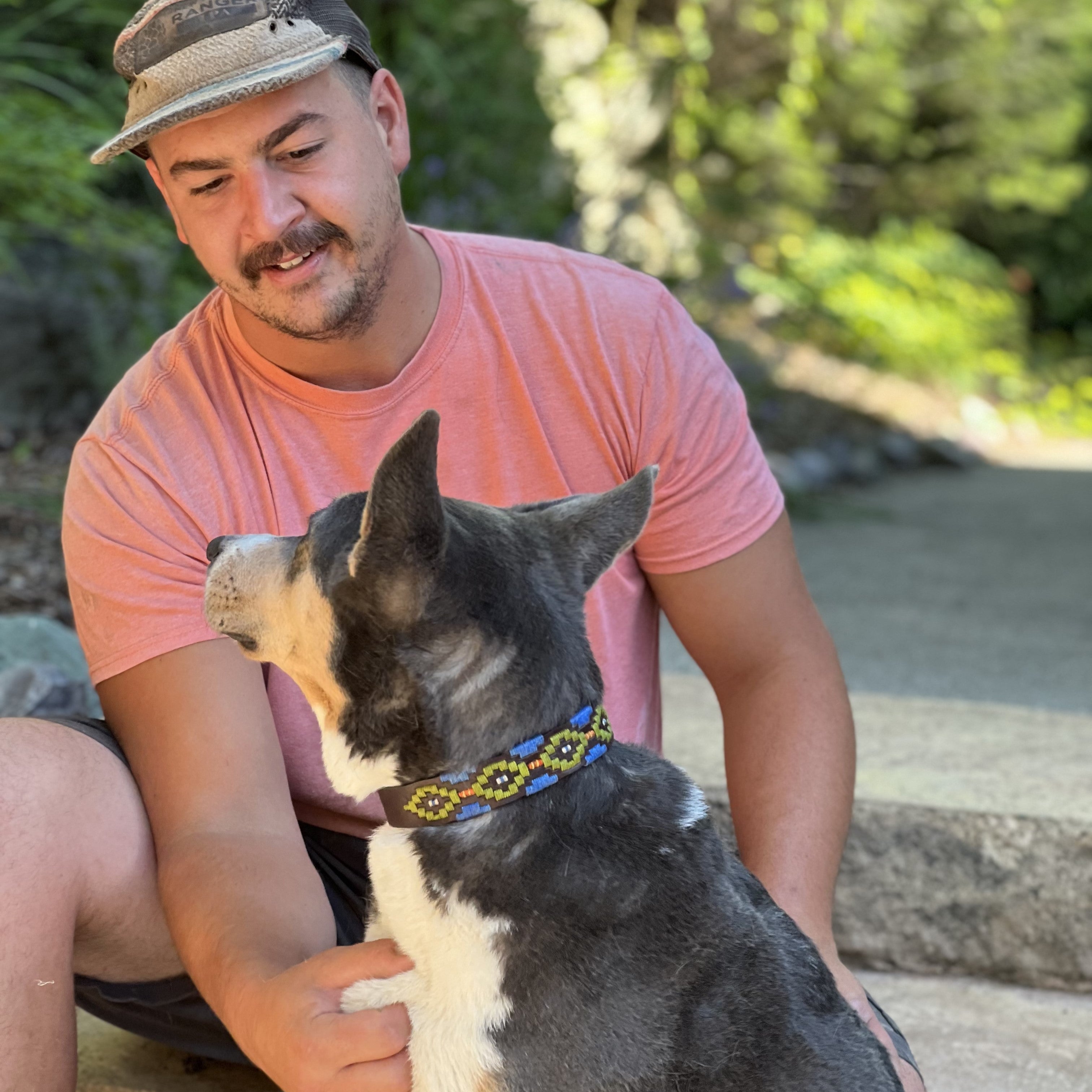 A man in a peach-colored t-shirt and a hat enjoys a moment with his loyal black and white dog, who sports a Georgie Paws sustainable Polo Collar - Albert, on a sunlit outdoor staircase surrounded by nature.