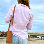 A person with curly hair, seen from the back, wears a pink shirt and light blue jeans, carrying a Georgie Paws Dog Walk Bag - Raw with brass hardware. They are on a beach with a cloudy sky.