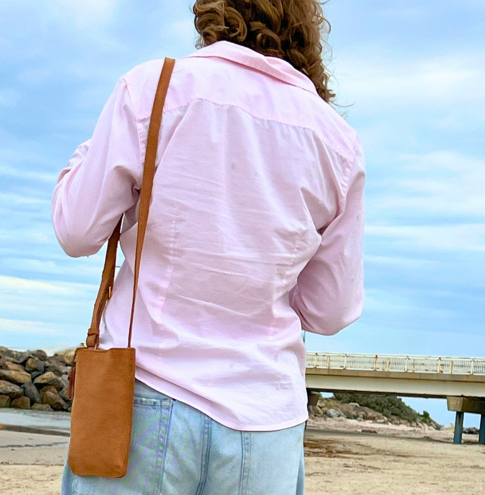 A person with curly hair seen from behind wearing a light pink shirt and light blue jeans, holding a Georgie Paws Dog Walk Bag - Grass, with a sandy beach and a pale sky in the background.