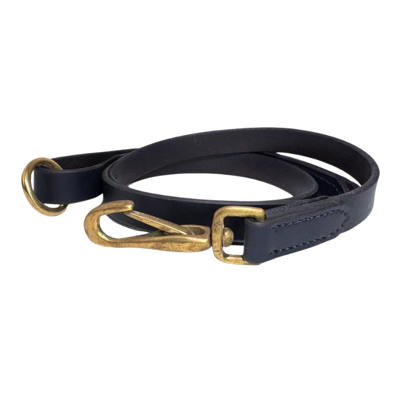 A coiled navy vegetable-tanned buffalo leather dog leash, the Bald Lead by Georgie Paws, with a brass clip lies on a white surface. The leash features a looped handle on one end and a metal D-ring near the handle for attaching accessories. This sustainable design ensures it is plastic-free.