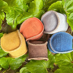 Five colorful, round suede jewelry pouches with snap closures, in earthy tones of brown, blue, yellow, gray, and red, nestled amongst lush green leaves and an eco-friendly Georgie Paws Brown Dog Ball.