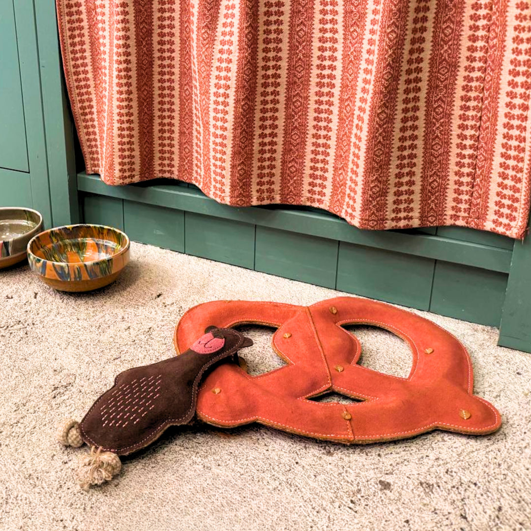 A Georgie Paws Baz the Bear Dog Toy in brown rests on a stone floor near a green cabinet, patterned ceramic bowl, metal water dish, and a red-and-cream patterned curtain in the background.