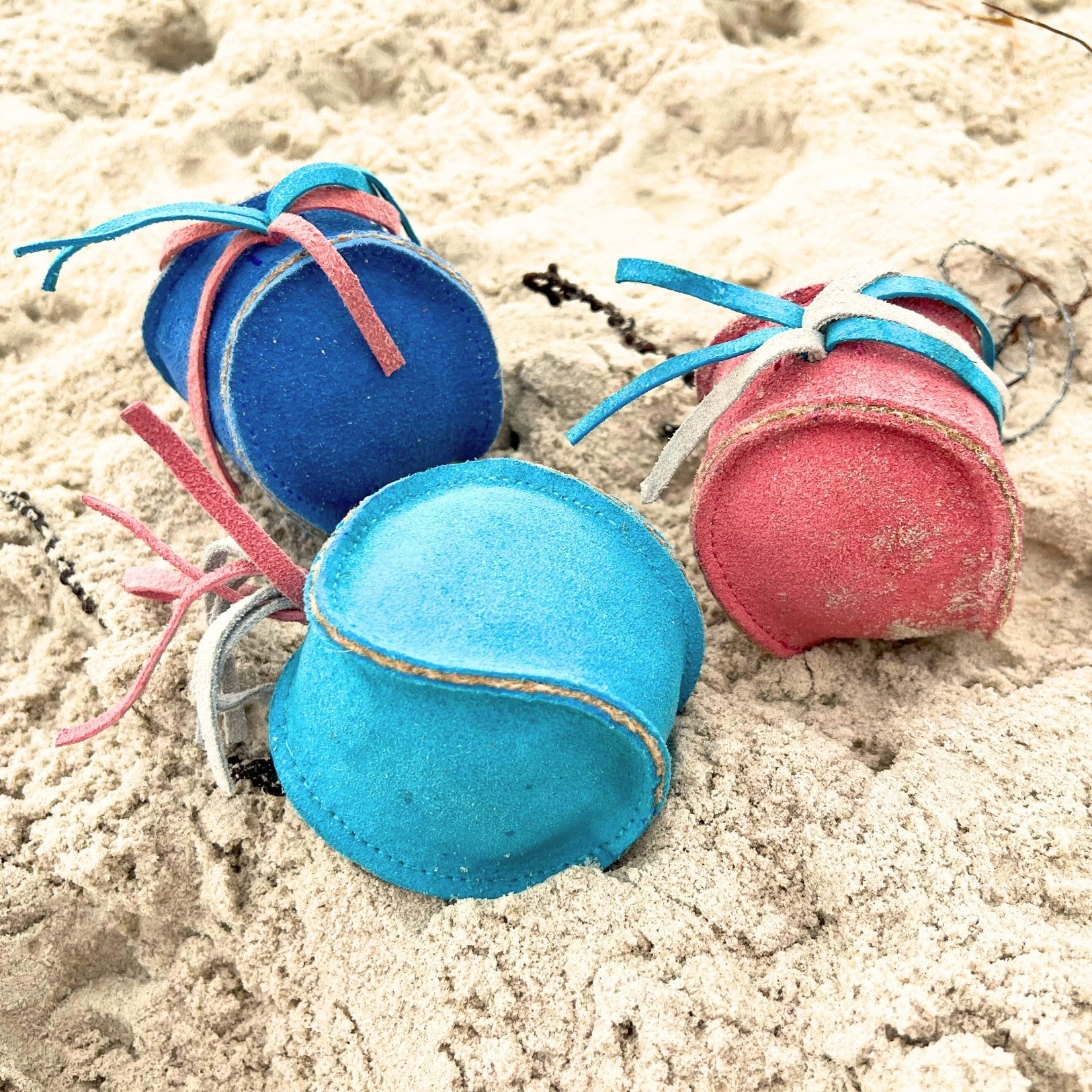Four colorful, worn-out Bobble Sea Urchin - blue juggling balls, made from buffalo suede and with shades of blue, pink, and red, rest on sandy ground, their laces partly buried, showing signs of frequent use.