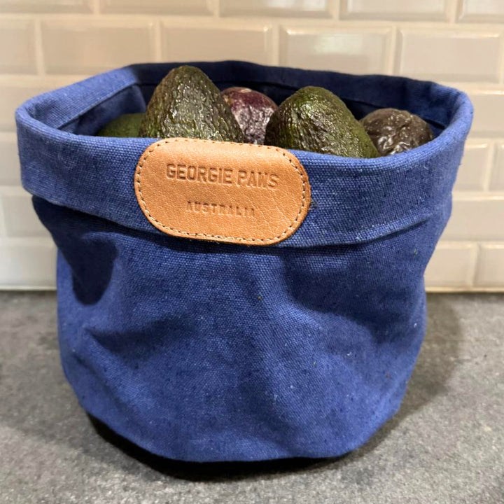 A blue Toy Storage Bucket from Georgie Paws, featuring a brown leather tag, holds several dark-skinned avocados on a grey countertop in front of a white subway tile backsplash.