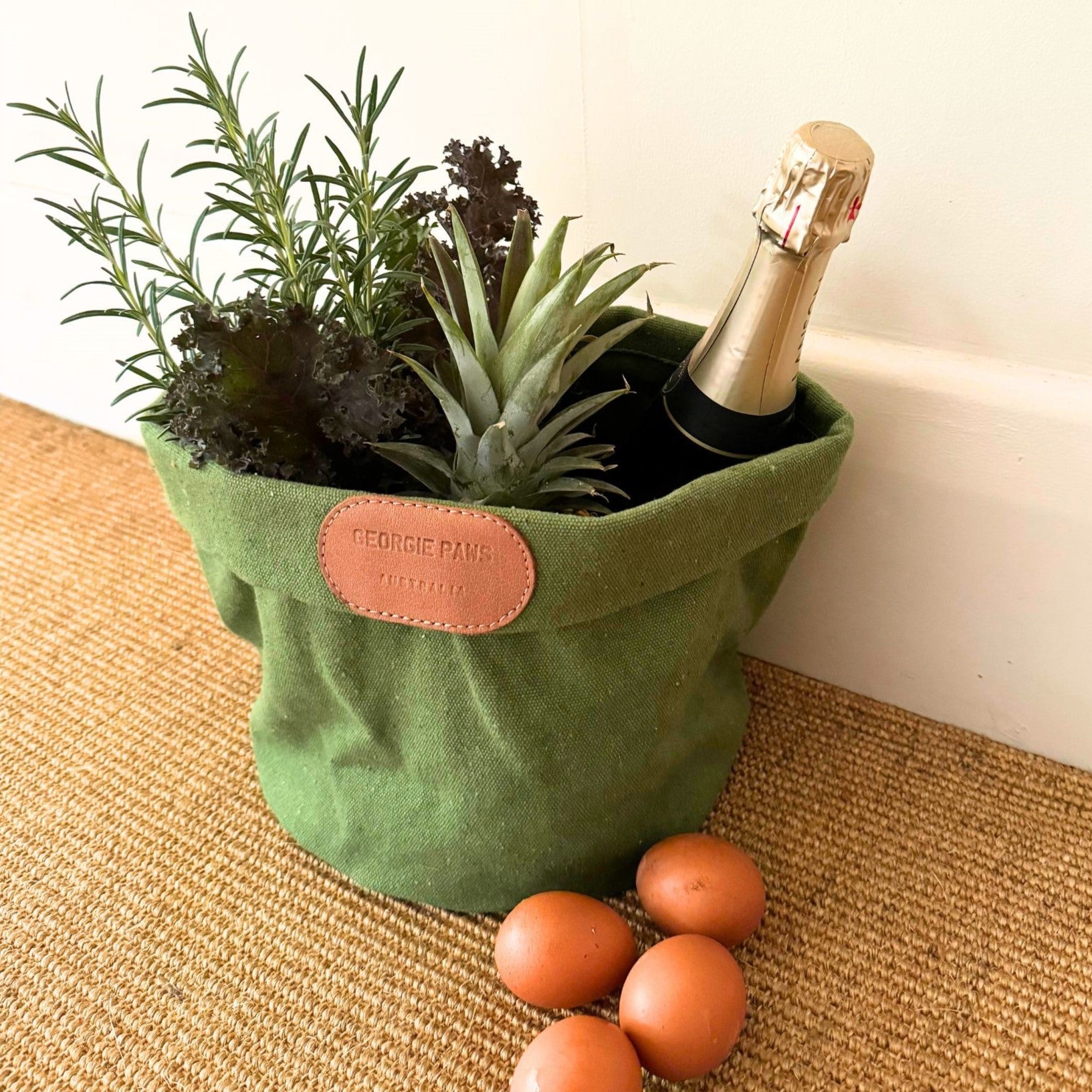 A green Toy Storage Bucket by Georgie Paws, crafted from heavy cotton canvas, holds an assortment of items: fresh rosemary, kale, a pineapple, and a bottle of champagne. The bucket has a leather label with "Georgie Paws" on it. Three brown eggs rest on the light brown woven carpet beside the versatile craft corner tote.