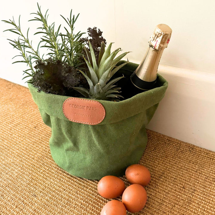 A green Toy Storage Bucket by Georgie Paws holds a pineapple, rosemary, leafy greens, and a champagne bottle. Four brown eggs sit on a beige woven mat in front of the bucket, which is placed near a white wall.