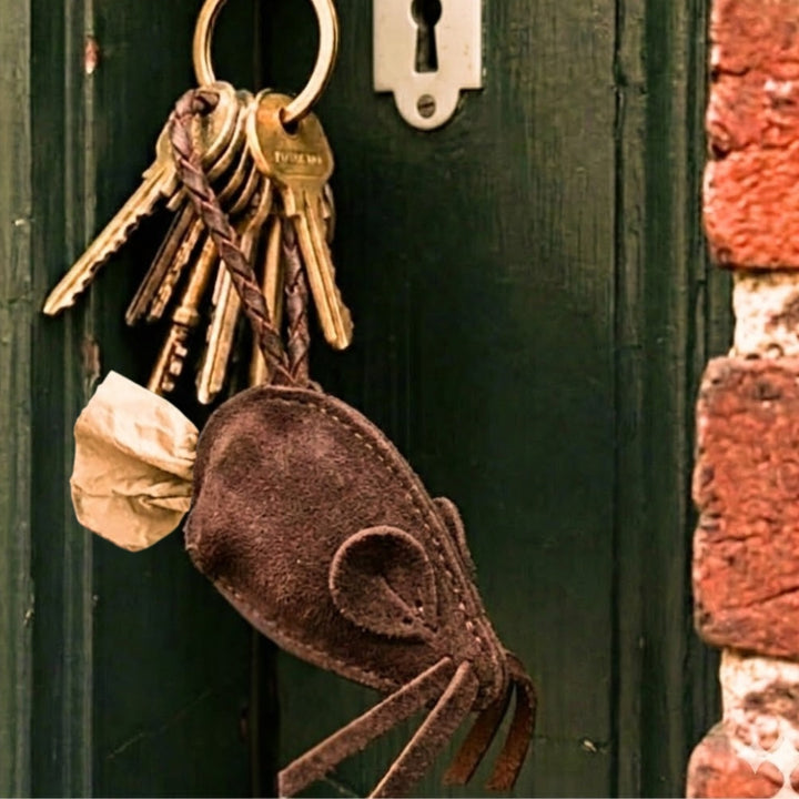 A cluster of keys hangs from a green door lock, featuring the Georgie Paws Mouse Poobag Dispenser in Carob—an eco-friendly dog accessory shaped like a mouse with a paper “tail”—set against a red brick wall.