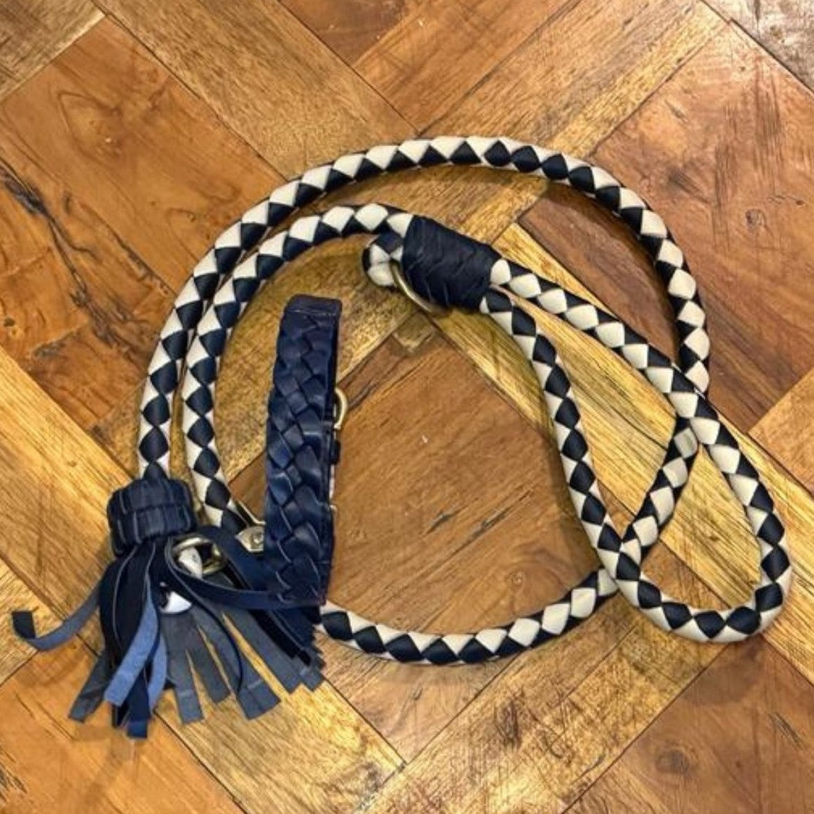 A navy Casey Collar dog leash by Georgie Paws, crafted from veg-tanned buffalo leather with a tasseled end and brass hardware, is coiled on a wooden parquet floor with rich brown tones and visible grain patterns.