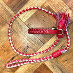 A braided red and white dog leash with a matching Georgie Paws Casey Collar - Red, featuring brass hardware and pink-red tassels, lies coiled on a parquet wood floor. The leash has a looped handle and metal ring.