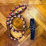 The Georgie Paws Casey Collar - Navy, a braided multicolored dog leash with a brown veg-tanned buffalo leather handle and brass hardware, is coiled on a diagonally patterned parquet floor beside a dark blue braided leather wrist strap.