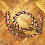 A braided dog leash with red, yellow, black, and tan strands lies coiled on a wooden floor. It features leather accents and brass hardware, ready to attach to the Georgie Paws Casey Collar - Syrup.
