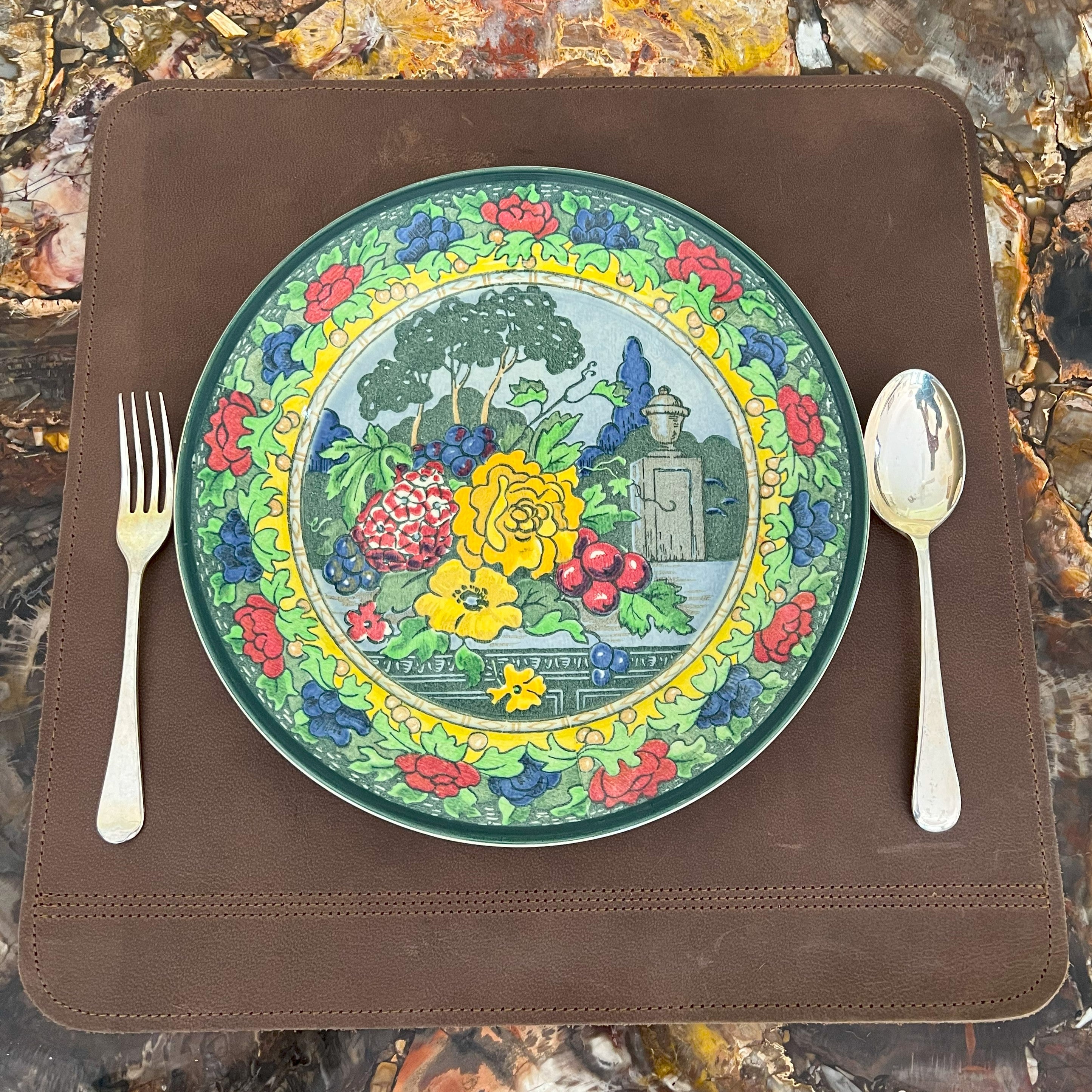 A decorative plate with vibrant floral and Hebrew text designs is set on a LIMITED EDITION buffalo leather Dinner Mat - classic - set of 2, flanked by a fork on the left and a spoon on the right, on a rustic table.