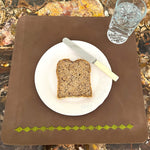 A slice of multigrain bread on a white plate with a knife on the right, set on LIMITED EDITION Buffalo Leather Dinner Mats embroidered with green patterns. Beside it, a clear glass of Georgie Paws Dinner Mat - grass - set of 2.