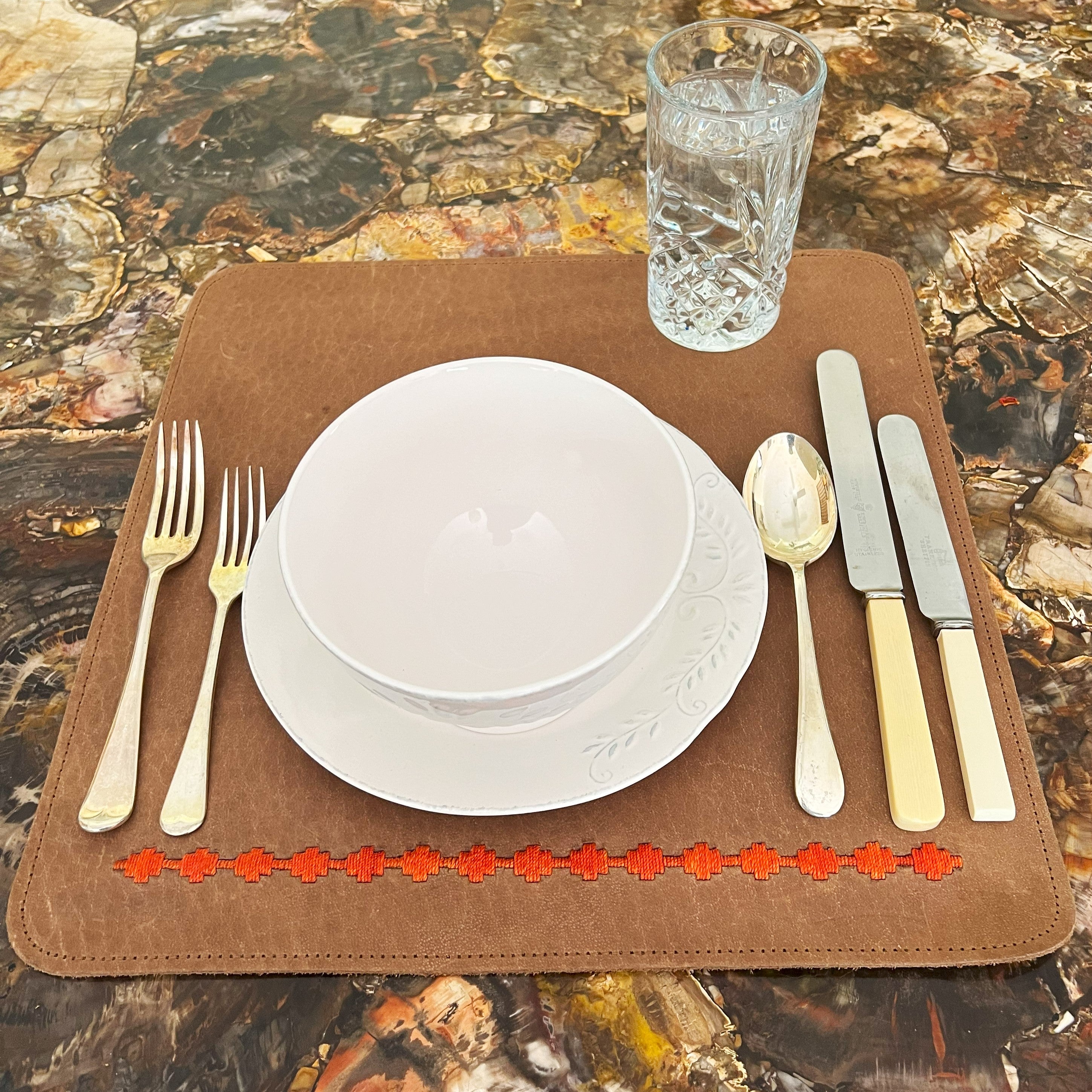 A table setting featuring a white plate with an intricate design on a LIMITED EDITION Georgie Paws buffalo leather dinner mat in ochre, adorned with a red floral pattern. Accompanied by two forks, a spoon, two knives.