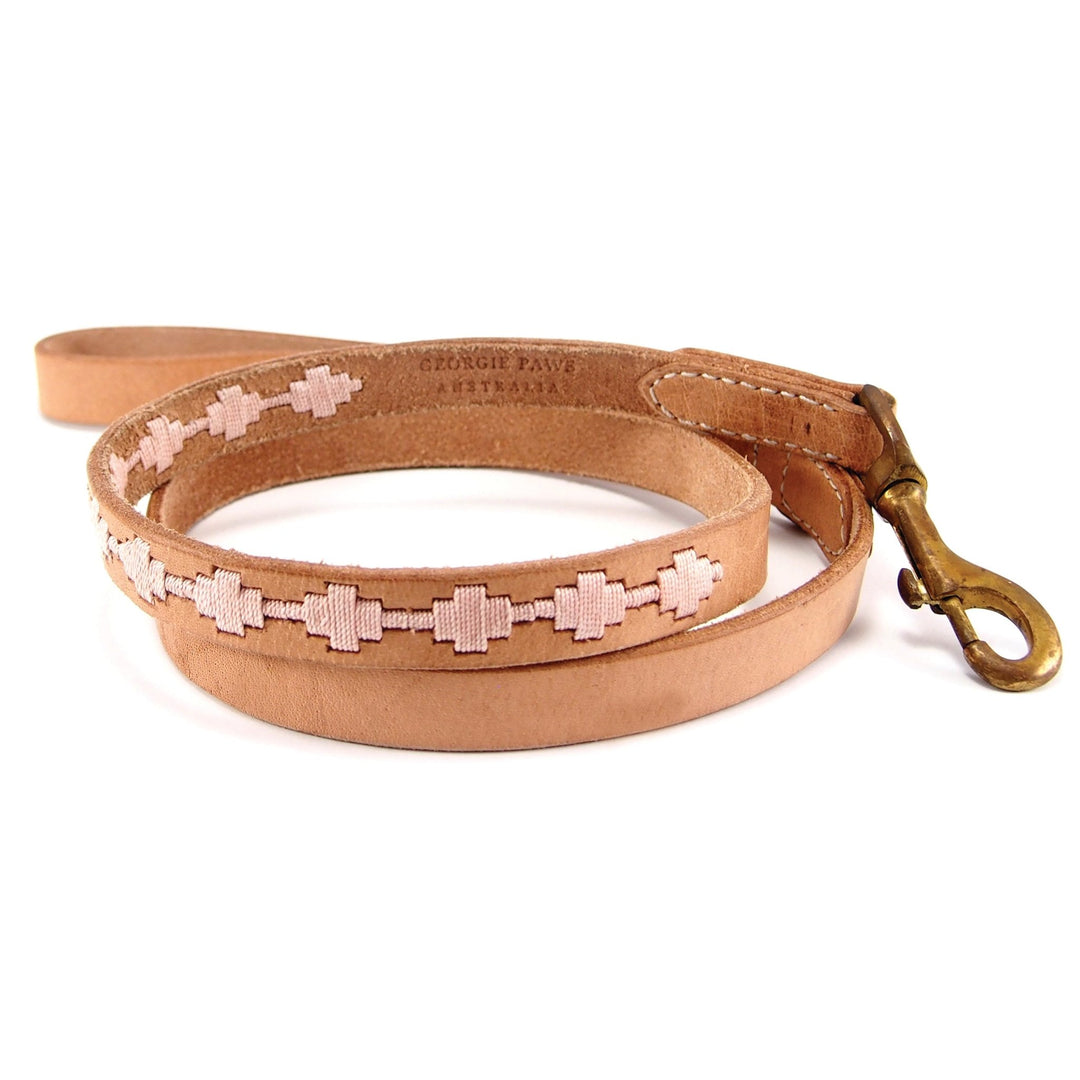 The Ethel Lead - Carnation by Georgie Paws is a tan, vegetable-tanned buffalo leather dog leash with a brass clasp, adorned along its length with pink geometric embroidery. Handmade and designed with a looped handle, the smooth, slightly shiny leather features the Georgie Paws logo embossed near the handle.