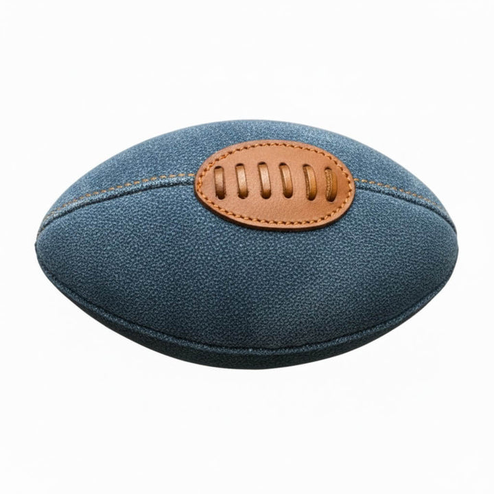 The Footy Dog Toy - Blue by Georgie Paws is a blue denim dog football toy featuring brown leather laces and a leather patch, displayed against a white background. The detailed stitching and textures of the denim and leather are clearly visible, highlighting its stylish design and eco-friendly appeal for your dog's playtime.