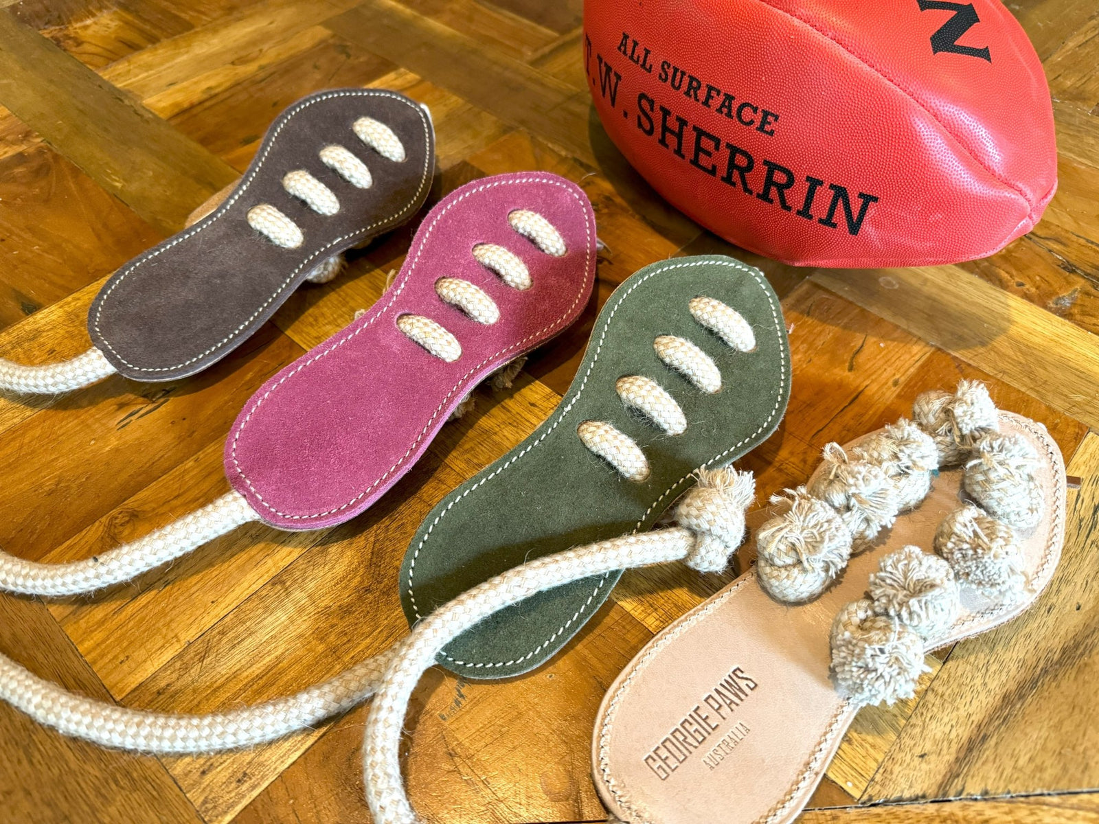 Three sustainable, sandal-shaped suede dog toys—the "Footy Boot" by Georgie Paws in hot pink, purple, and green—each featuring rope laces, rest on a wooden surface beside a red Sherrin Australian football. One natural-fiber toy displays the "GEORGIE PAWS AUSTRALIA" branding and is accented with knotted rope details.