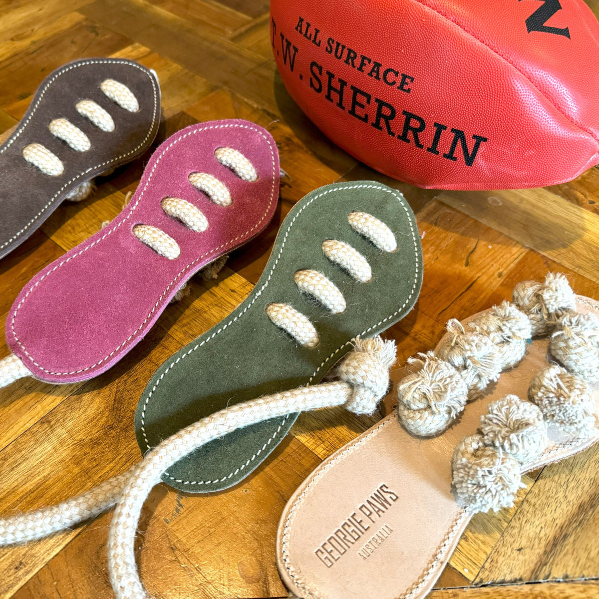 Three Georgie Paws Footy Boot suede dog tug toys in purple, green and tan with rope laces, next to a red Sherrin football. The tan toy is stamped GEORGIE PAWS AUSTRALIA.