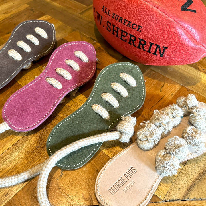 Three Georgie Paws Footy Boot suede dog tug toys in purple, green and tan with rope laces, next to a red Sherrin football. The tan toy is stamped GEORGIE PAWS AUSTRALIA.