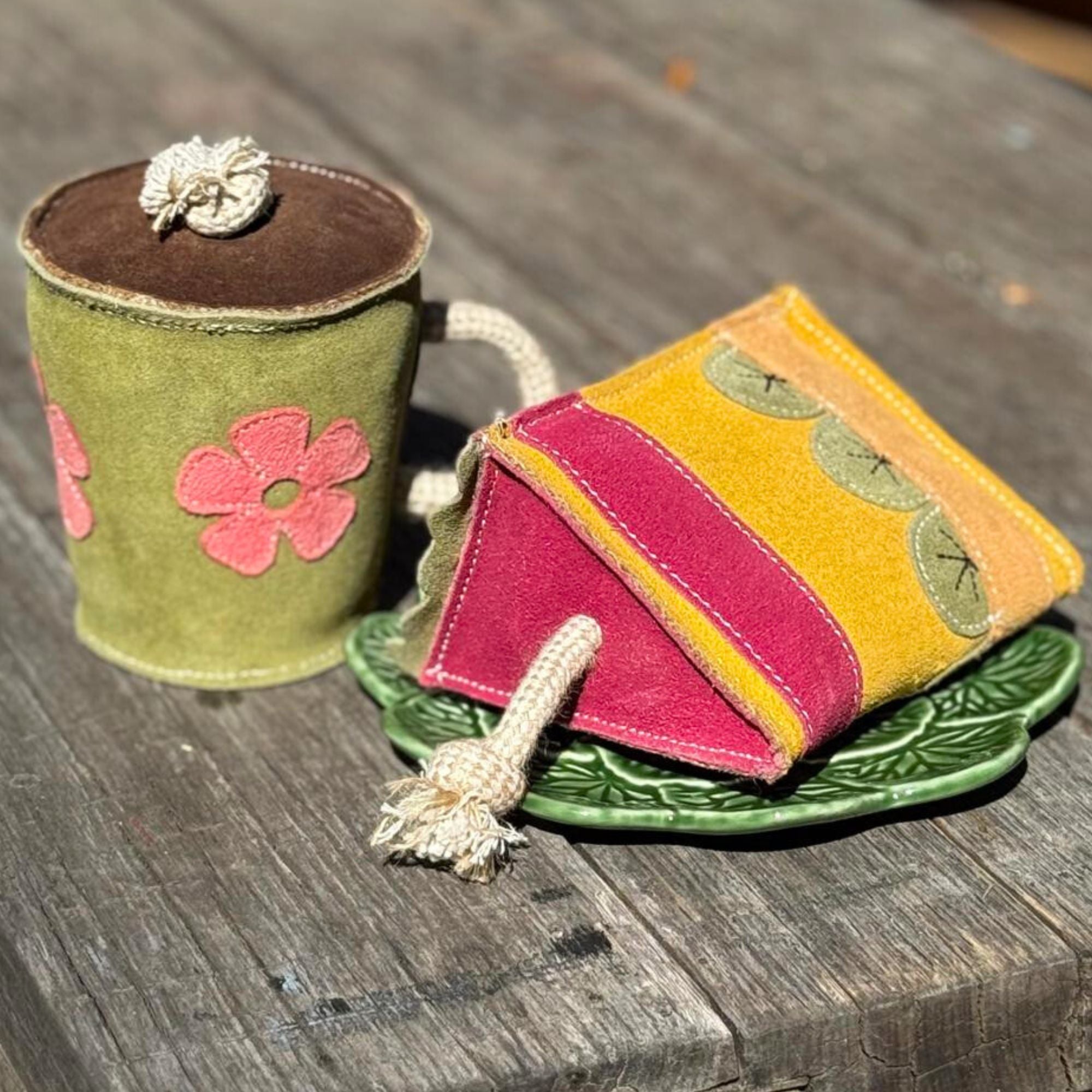 A whimsical fabric-crafted teacup and teabag rest on a green leaf-patterned dish, evoking the Georgie Paws Hotto Chocco Toy. The green cup features pink flowers and a rope handle that beautifully match the colorful teabag's pink, yellow, and green patches against the wooden surface.