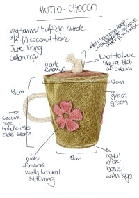 The Hotto Chocco Dog Toy by Georgie Paws is a hand-drawn tan cup with "HOTTO-CHOCKO" notes, pink flower accents, a green base, dark brown jute lining, rope handle, and detailed stitching.
