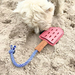 A fluffy white dog curiously sniffs at a Georgie Paws Icy Pole - pink dog toy partially buried in the sand, designed for teething puppies and featuring a buffalo leather and suede blue and white rope.
