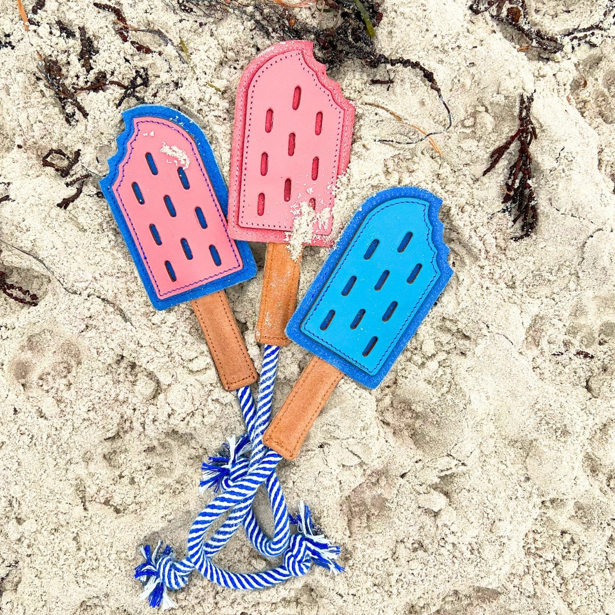 Three colorful compostable dog toy decorations with Georgie Paws Icy Pole - blue tops, along with brown sticks and a blue-and-white striped ribbon, are placed on sand, suggesting a whimsical summer beach theme.