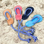 Three pairs of colorful buffalo leather Georgie Paws blue Jandal flip-flops arranged in a circle, connected by a blue and white striped rope, lie on a sandy beach, depicting a leisurely, summer vibe.