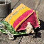 A vibrant fabric Kiwi Sponge Cake tea cozy by Georgie Paws, with yellow and red panels and a rope "cherry" topper, sits on a green leaf-patterned plate. Beside it is a decorative silver fork, while a cup of black coffee rests in the background on a wooden surface.