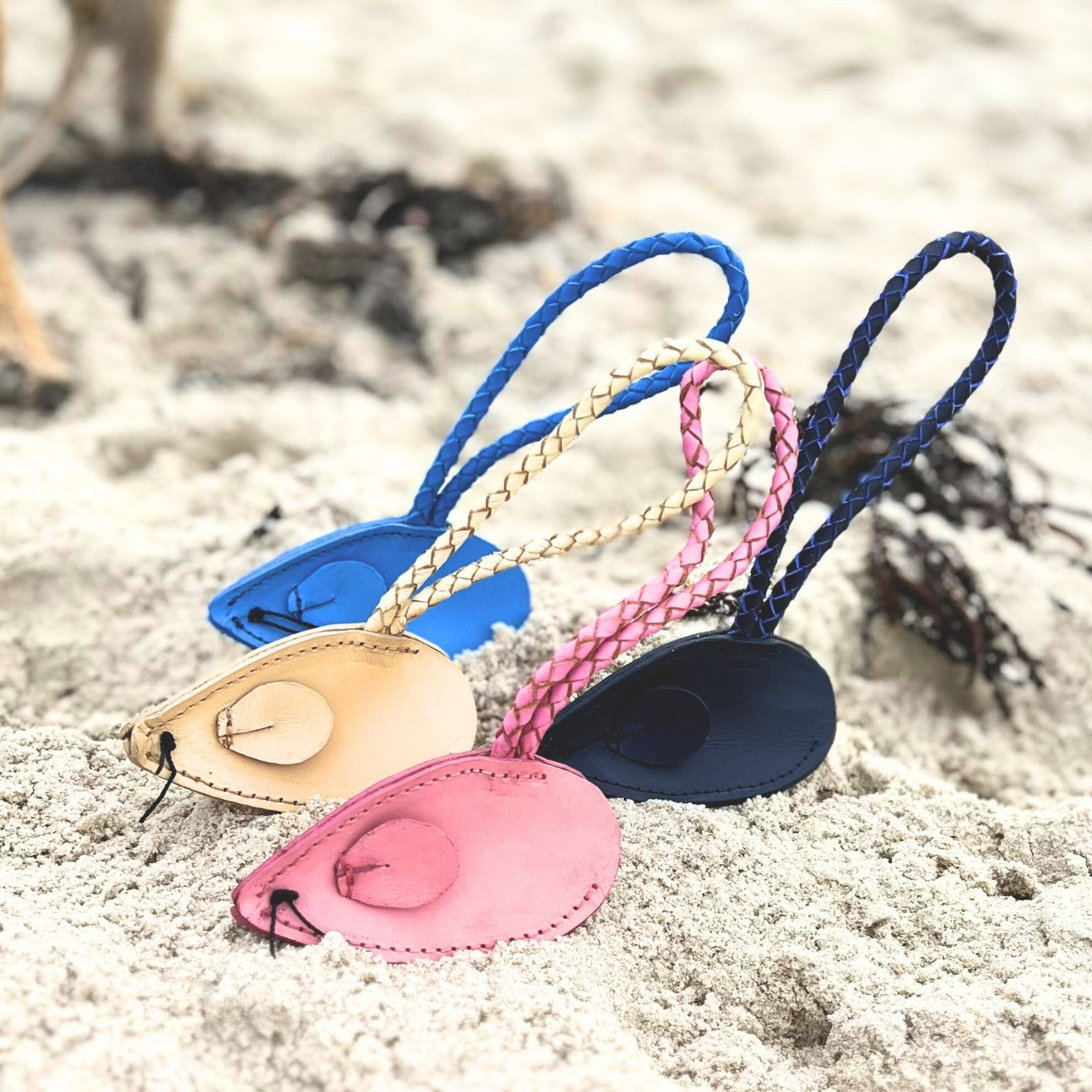 Four colorful Georgie Paws buffalo leather flip-flops with braided straps are playfully stuck upright in sandy beach terrain, evoking a laid-back, beachy vibe.