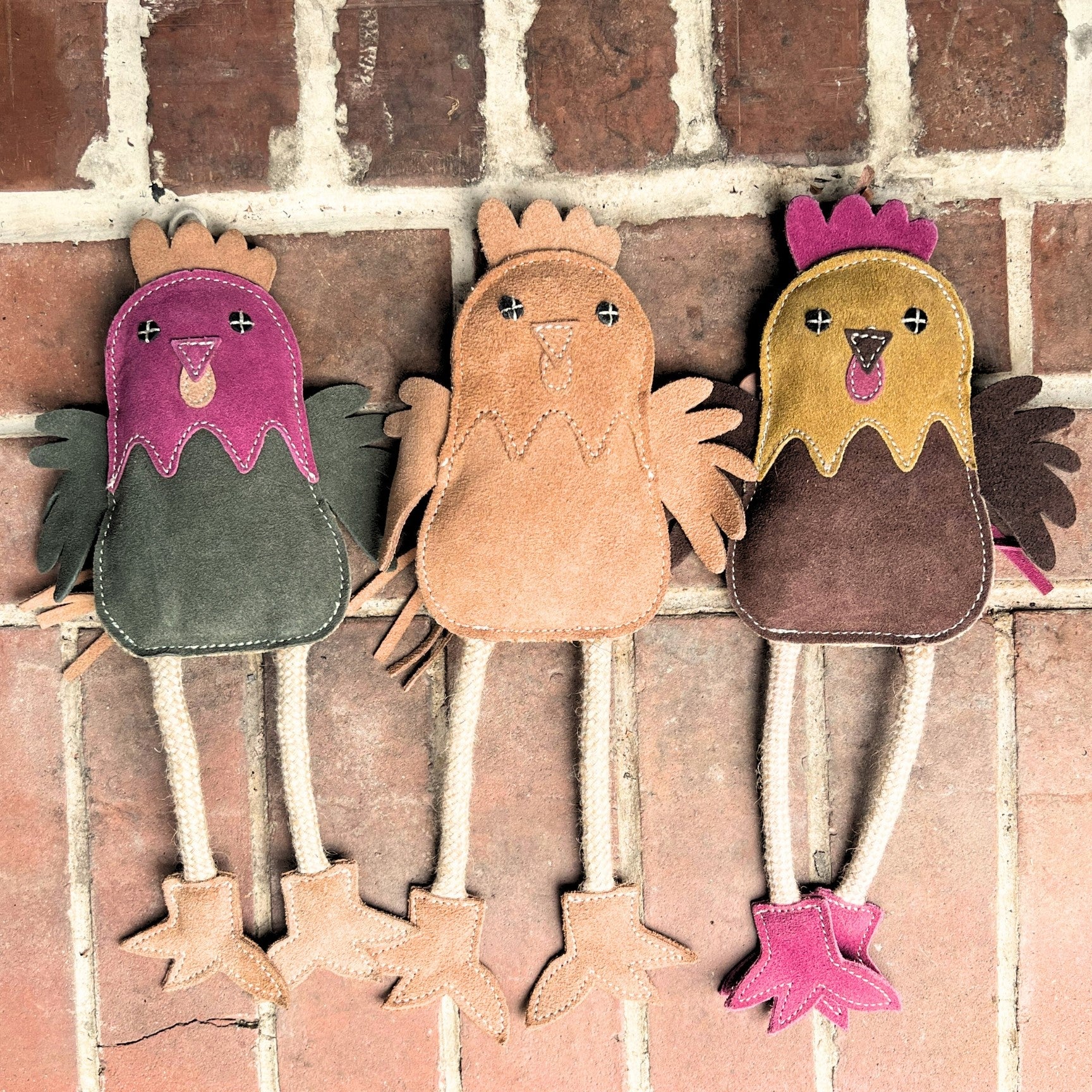 Three handmade, colorful Matilda the Chicken plushies from Georgie Paws are lined up against a brick background. Each Matilda plushie, sporting a unique color scheme—green and purple, brown and pink, and yellow and brown—could double as a sustainable dog toy. The chickens feature long rope-like legs, felt wings, and stitched facial features.