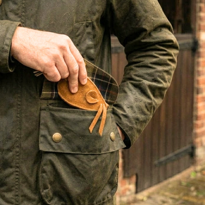 A person in a green waxed jacket with brass snap buttons and plaid lining puts a Georgie Paws Mouse Poobag Dispenser - Sand into their large front pocket. A brick wall and wooden door appear blurred in the background.
