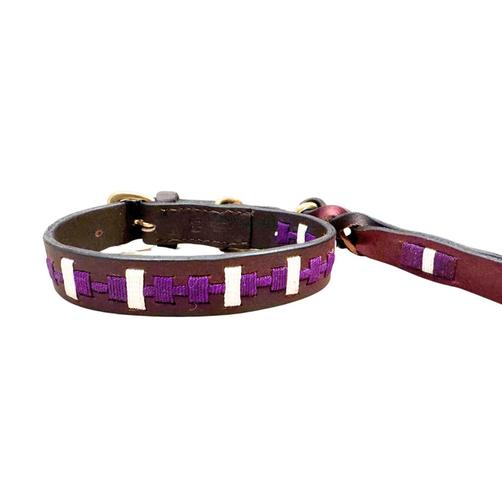 Introducing the Polo Collar - Lokki Purple from Georgie Paws—a brown leather dog collar adorned with purple and white ribbon woven in a rectangular pattern along its length. Featuring sturdy metal hardware for secure leash attachment, this design is both simple and stylish, providing a sustainable choice for your pet.