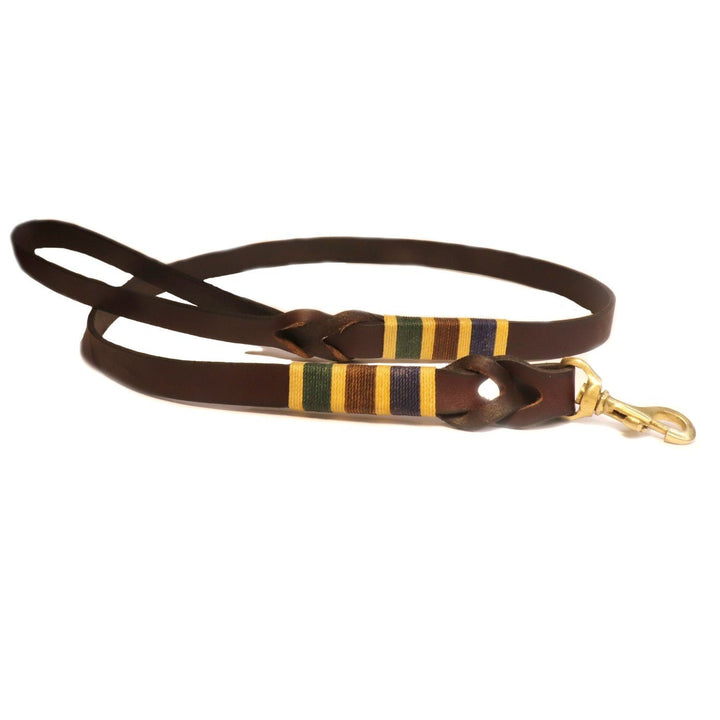 The Polo Lead - Jinky by Georgie Paws is a brown, natural vegetable tanned buffalo leather dog leash with a loop handle and gold metal clasp. Decorative yellow, green, and purple thread is wrapped in three bands near the clasp, adding a colorful accent. The smooth leather leash is shown laid flat in a loose oval shape.